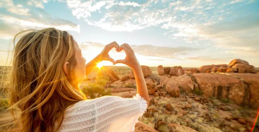 Junge Frau formt ein Herz mit ihren Händen vor dem Sonnenuntergang im Marbles Conservation Reserve, Australien
